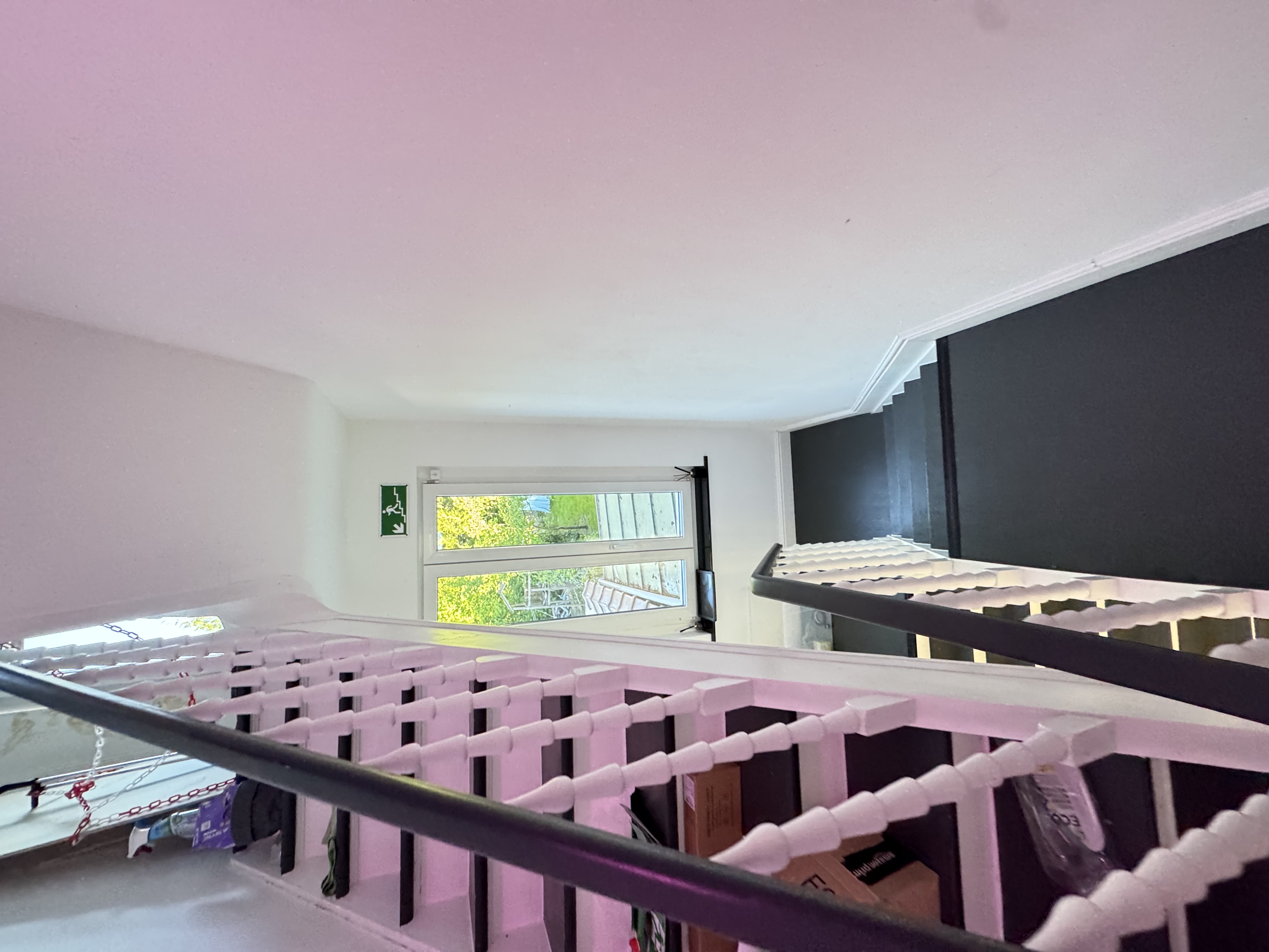Stairwell with green Exit pictogram pointing downward, white-painted spindle balustrade, daylight window at the half-landing.